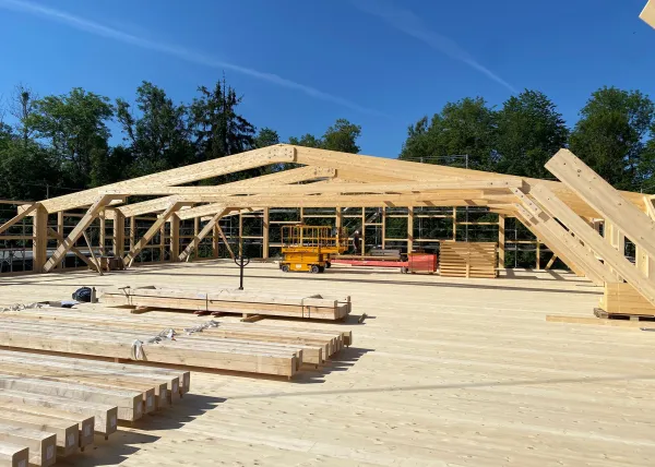 Innenansicht einer im Bau befindlichen Gewerbehalle mit weit gespannter Holzdachkonstruktion aus Brettschichtholz. Sichtbar sind die tragenden Binder, Sparren und die Dachschalung, während eine Hebebühne auf der Holzdecke den Baufortschritt der Umnutzung dokumentiert.