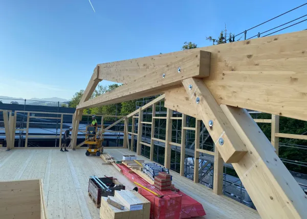 Innenansicht einer im Bau befindlichen Gewerbehalle mit weit gespannter Holzdachkonstruktion aus Brettschichtholz. Sichtbar sind die tragenden Binder, Sparren und die Dachschalung, während eine Hebebühne auf der Holzdecke den Baufortschritt der Umnutzung dokumentiert.