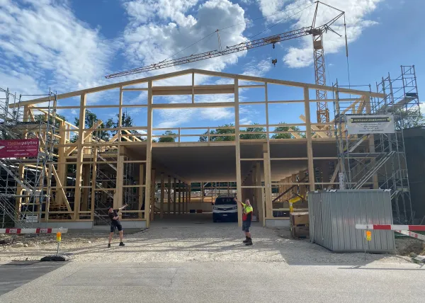 Innenansicht einer im Bau befindlichen Gewerbehalle mit weit gespannter Holzdachkonstruktion aus Brettschichtholz. Sichtbar sind die tragenden Binder, Sparren und die Dachschalung, während eine Hebebühne auf der Holzdecke den Baufortschritt der Umnutzung dokumentiert.
