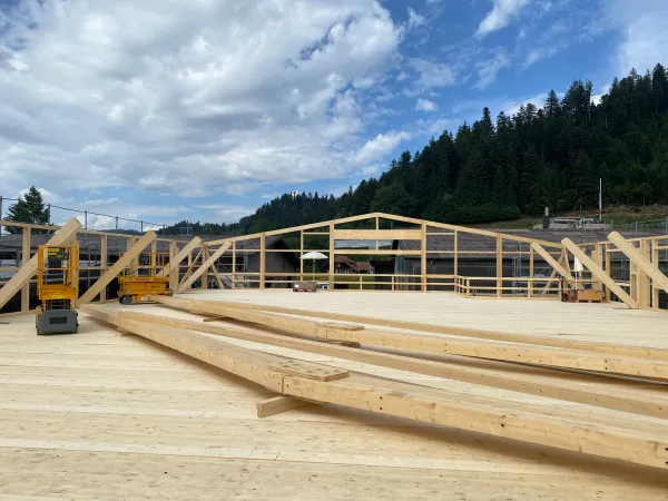 Innenansicht einer im Bau befindlichen Gewerbehalle mit weit gespannter Holzdachkonstruktion aus Brettschichtholz. Sichtbar sind die tragenden Binder, Sparren und die Dachschalung, während eine Hebebühne auf der Holzdecke den Baufortschritt der Umnutzung dokumentiert.