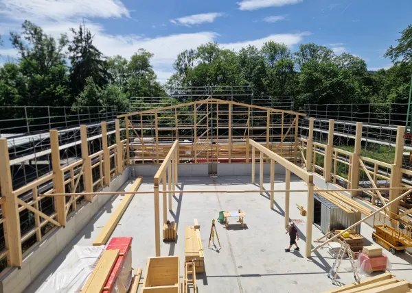 Innenansicht einer im Bau befindlichen Gewerbehalle mit weit gespannter Holzdachkonstruktion aus Brettschichtholz. Sichtbar sind die tragenden Binder, Sparren und die Dachschalung, während eine Hebebühne auf der Holzdecke den Baufortschritt der Umnutzung dokumentiert.