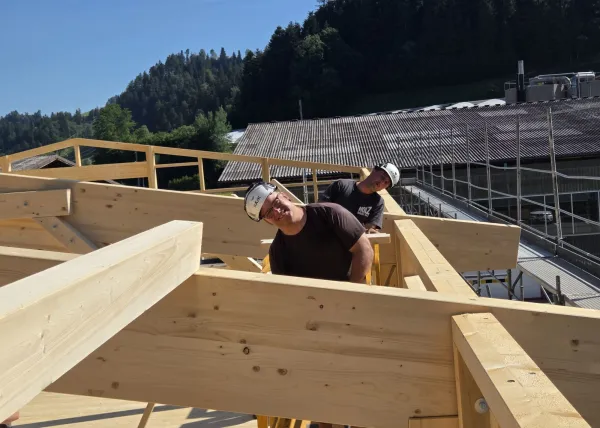 Innenansicht einer im Bau befindlichen Gewerbehalle mit weit gespannter Holzdachkonstruktion aus Brettschichtholz. Sichtbar sind die tragenden Binder, Sparren und die Dachschalung, während eine Hebebühne auf der Holzdecke den Baufortschritt der Umnutzung dokumentiert.