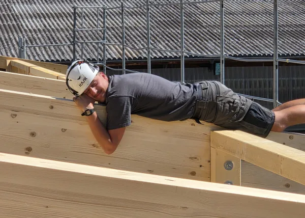 Innenansicht einer im Bau befindlichen Gewerbehalle mit weit gespannter Holzdachkonstruktion aus Brettschichtholz. Sichtbar sind die tragenden Binder, Sparren und die Dachschalung, während eine Hebebühne auf der Holzdecke den Baufortschritt der Umnutzung dokumentiert.