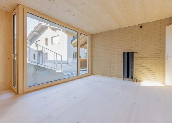 einblicke in ein holzbauprojekt in gunten: die aussenansicht zeigt ein haus mit heller holzfassade und balkon, eingebettet zwischen bäumen und einem steinmäuerchen. im inneren prägen helle holzdecken und sichtbare balken, grosse fenster und warme holzoberflächen die räume. zu sehen sind ein grosszügiger wohnbereich mit zugang zum balkon und ausblick ins grüne, eine holzküche mit arbeitsinsel sowie ein badezimmer mit rundem spiegel und aufgesetztem waschbecken. helles ziegelmauerwerk und sorgfältig erhaltene