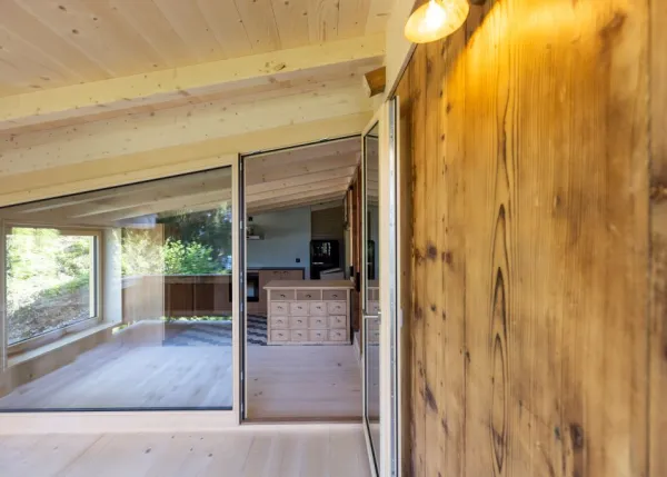 einblicke in ein holzbauprojekt in gunten: die aussenansicht zeigt ein haus mit heller holzfassade und balkon, eingebettet zwischen bäumen und einem steinmäuerchen. im inneren prägen helle holzdecken und sichtbare balken, grosse fenster und warme holzoberflächen die räume. zu sehen sind ein grosszügiger wohnbereich mit zugang zum balkon und ausblick ins grüne, eine holzküche mit arbeitsinsel sowie ein badezimmer mit rundem spiegel und aufgesetztem waschbecken. helles ziegelmauerwerk und sorgfältig erhaltene