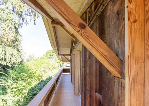 einblicke in ein holzbauprojekt in gunten: die aussenansicht zeigt ein haus mit heller holzfassade und balkon, eingebettet zwischen bäumen und einem steinmäuerchen. im inneren prägen helle holzdecken und sichtbare balken, grosse fenster und warme holzoberflächen die räume. zu sehen sind ein grosszügiger wohnbereich mit zugang zum balkon und ausblick ins grüne, eine holzküche mit arbeitsinsel sowie ein badezimmer mit rundem spiegel und aufgesetztem waschbecken. helles ziegelmauerwerk und sorgfältig erhaltene