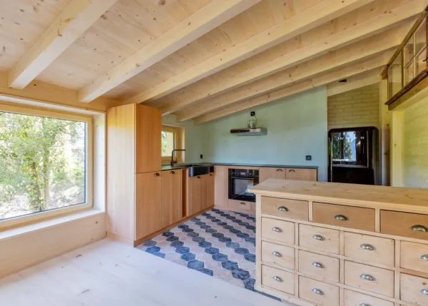 einblicke in ein holzbauprojekt in gunten: die aussenansicht zeigt ein haus mit heller holzfassade und balkon, eingebettet zwischen bäumen und einem steinmäuerchen. im inneren prägen helle holzdecken und sichtbare balken, grosse fenster und warme holzoberflächen die räume. zu sehen sind ein grosszügiger wohnbereich mit zugang zum balkon und ausblick ins grüne, eine holzküche mit arbeitsinsel sowie ein badezimmer mit rundem spiegel und aufgesetztem waschbecken. helles ziegelmauerwerk und sorgfältig erhaltene