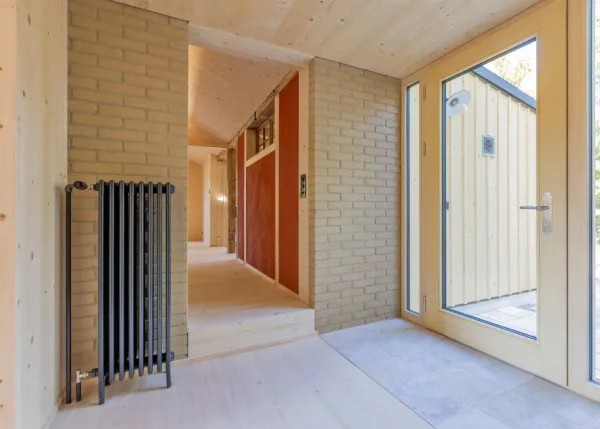 einblicke in ein holzbauprojekt in gunten: die aussenansicht zeigt ein haus mit heller holzfassade und balkon, eingebettet zwischen bäumen und einem steinmäuerchen. im inneren prägen helle holzdecken und sichtbare balken, grosse fenster und warme holzoberflächen die räume. zu sehen sind ein grosszügiger wohnbereich mit zugang zum balkon und ausblick ins grüne, eine holzküche mit arbeitsinsel sowie ein badezimmer mit rundem spiegel und aufgesetztem waschbecken. helles ziegelmauerwerk und sorgfältig erhaltene