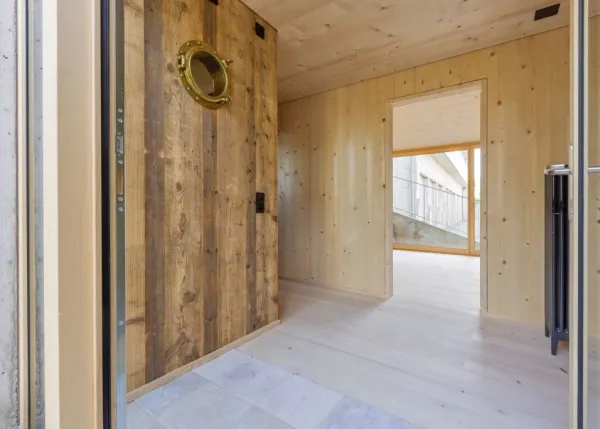 einblicke in ein holzbauprojekt in gunten: die aussenansicht zeigt ein haus mit heller holzfassade und balkon, eingebettet zwischen bäumen und einem steinmäuerchen. im inneren prägen helle holzdecken und sichtbare balken, grosse fenster und warme holzoberflächen die räume. zu sehen sind ein grosszügiger wohnbereich mit zugang zum balkon und ausblick ins grüne, eine holzküche mit arbeitsinsel sowie ein badezimmer mit rundem spiegel und aufgesetztem waschbecken. helles ziegelmauerwerk und sorgfältig erhaltene