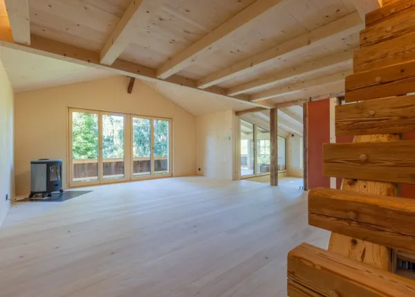 einblicke in ein holzbauprojekt in gunten: die aussenansicht zeigt ein haus mit heller holzfassade und balkon, eingebettet zwischen bäumen und einem steinmäuerchen. im inneren prägen helle holzdecken und sichtbare balken, grosse fenster und warme holzoberflächen die räume. zu sehen sind ein grosszügiger wohnbereich mit zugang zum balkon und ausblick ins grüne, eine holzküche mit arbeitsinsel sowie ein badezimmer mit rundem spiegel und aufgesetztem waschbecken. helles ziegelmauerwerk und sorgfältig erhaltene