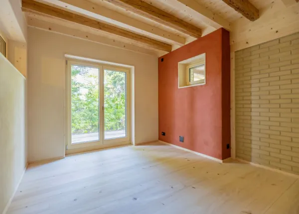 einblicke in ein holzbauprojekt in gunten: die aussenansicht zeigt ein haus mit heller holzfassade und balkon, eingebettet zwischen bäumen und einem steinmäuerchen. im inneren prägen helle holzdecken und sichtbare balken, grosse fenster und warme holzoberflächen die räume. zu sehen sind ein grosszügiger wohnbereich mit zugang zum balkon und ausblick ins grüne, eine holzküche mit arbeitsinsel sowie ein badezimmer mit rundem spiegel und aufgesetztem waschbecken. helles ziegelmauerwerk und sorgfältig erhaltene