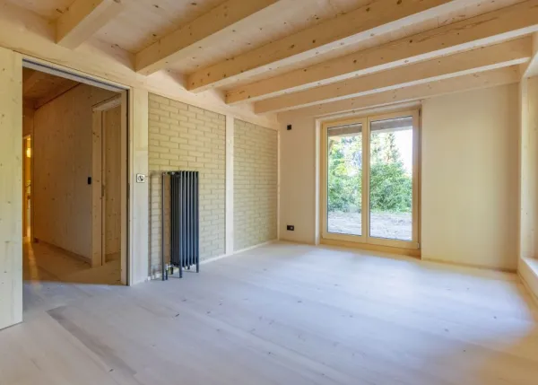einblicke in ein holzbauprojekt in gunten: die aussenansicht zeigt ein haus mit heller holzfassade und balkon, eingebettet zwischen bäumen und einem steinmäuerchen. im inneren prägen helle holzdecken und sichtbare balken, grosse fenster und warme holzoberflächen die räume. zu sehen sind ein grosszügiger wohnbereich mit zugang zum balkon und ausblick ins grüne, eine holzküche mit arbeitsinsel sowie ein badezimmer mit rundem spiegel und aufgesetztem waschbecken. helles ziegelmauerwerk und sorgfältig erhaltene