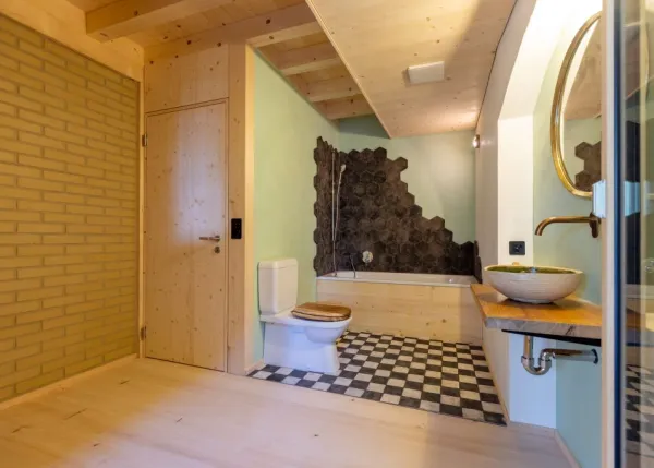 einblicke in ein holzbauprojekt in gunten: die aussenansicht zeigt ein haus mit heller holzfassade und balkon, eingebettet zwischen bäumen und einem steinmäuerchen. im inneren prägen helle holzdecken und sichtbare balken, grosse fenster und warme holzoberflächen die räume. zu sehen sind ein grosszügiger wohnbereich mit zugang zum balkon und ausblick ins grüne, eine holzküche mit arbeitsinsel sowie ein badezimmer mit rundem spiegel und aufgesetztem waschbecken. helles ziegelmauerwerk und sorgfältig erhaltene
