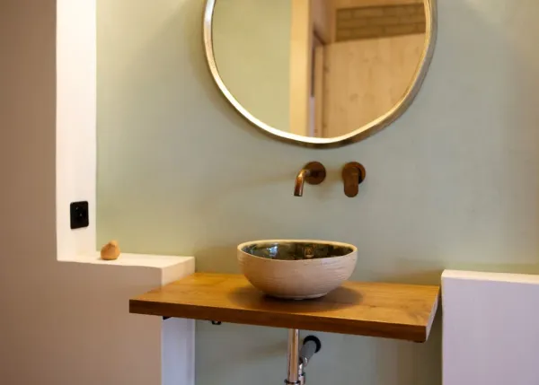 einblicke in ein holzbauprojekt in gunten: die aussenansicht zeigt ein haus mit heller holzfassade und balkon, eingebettet zwischen bäumen und einem steinmäuerchen. im inneren prägen helle holzdecken und sichtbare balken, grosse fenster und warme holzoberflächen die räume. zu sehen sind ein grosszügiger wohnbereich mit zugang zum balkon und ausblick ins grüne, eine holzküche mit arbeitsinsel sowie ein badezimmer mit rundem spiegel und aufgesetztem waschbecken. helles ziegelmauerwerk und sorgfältig erhaltene