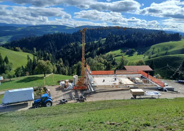 Holzbau im Rohbau mit Baukran an einem grünen Hang, mit bewaldeten Hügeln und schneebedeckten Bergen im Hintergrund.