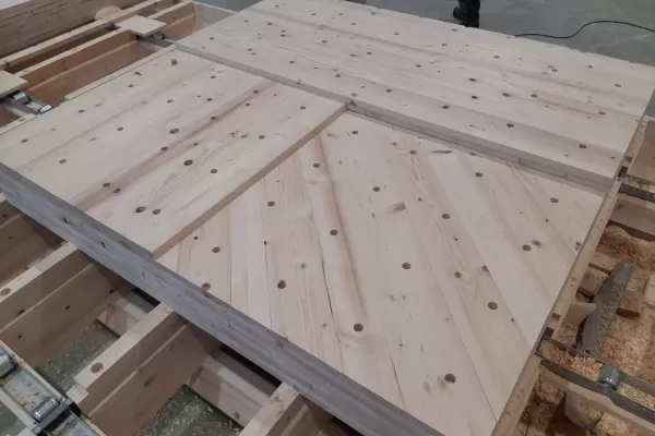 Produktionsstart Wandelemente Kunstpavillons   In unserer Produktionsstätte in Trub haben die Arbeiten zu den Wandelementen begonnen. Die Fertigungsanlage umfasst einen Produktionstisch, ein Dübelportal und ein Abbundportal. Die auf dem Produktionstisch ausgelegten Bretter werden vom Dübelportal vollautomatisch bei den Kreuzungspunkten verbunden. Das Abbundportal fräst die Konturen, bohrt die Steckdosen, kehlt die Verbindungsnuten und kalibriert die Elemente auf die gewünschte Dicke.