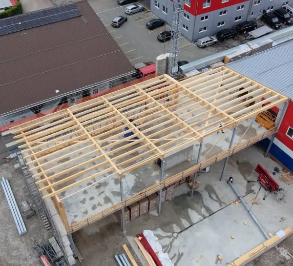 Luftaufnahme eines Gewerbebaus während der Bauphase. Zu sehen ist die im Aufbau befindliche Holzdachkonstruktion mit Tragwerk und Sparren auf einem Industriegebäude, umgeben von Werkhallen und Baustelleneinrichtungen.