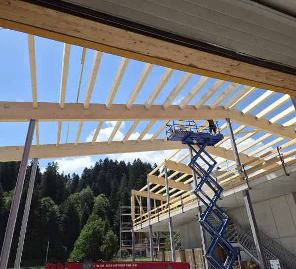 Luftaufnahme eines Gewerbebaus während der Bauphase. Zu sehen ist die im Aufbau befindliche Holzdachkonstruktion mit Tragwerk und Sparren auf einem Industriegebäude, umgeben von Werkhallen und Baustelleneinrichtungen.