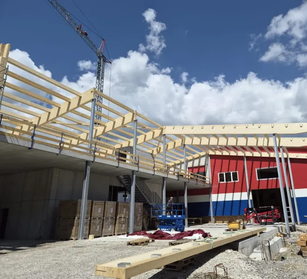 Luftaufnahme eines Gewerbebaus während der Bauphase. Zu sehen ist die im Aufbau befindliche Holzdachkonstruktion mit Tragwerk und Sparren auf einem Industriegebäude, umgeben von Werkhallen und Baustelleneinrichtungen.